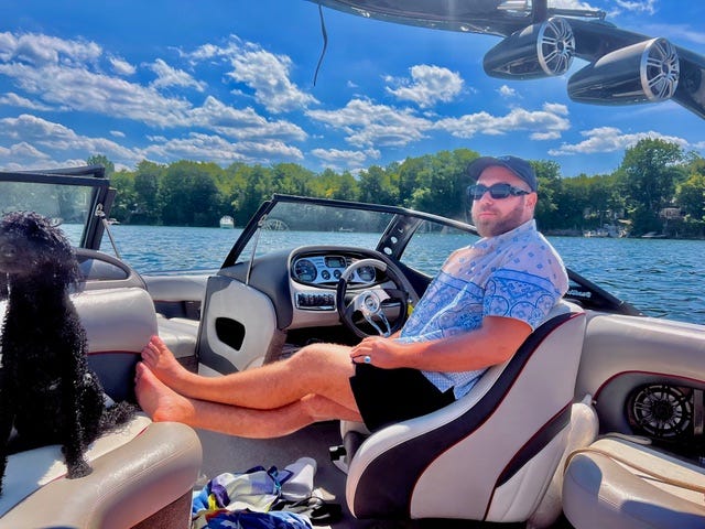 Person relaxing on a boat with a dog, surrounded by water and trees.