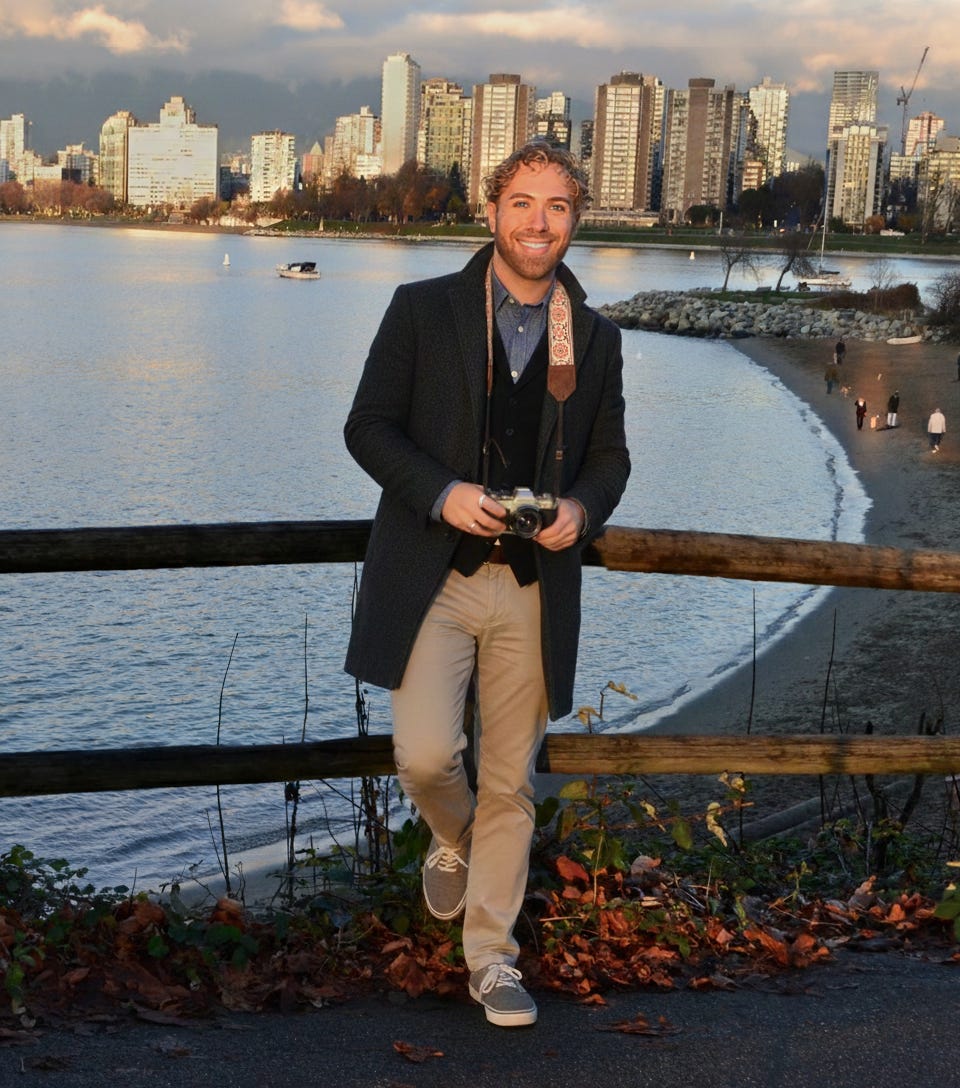 A person holding a camera near a waterfront with a city skyline in the background.