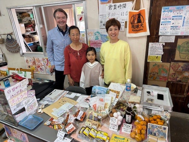A family standing behind a market stall filled with various food items and products.