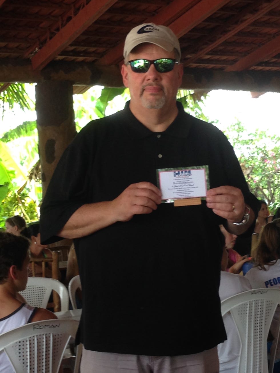 Glen Wagner in sunglasses and cap holding a small plaque indoors.