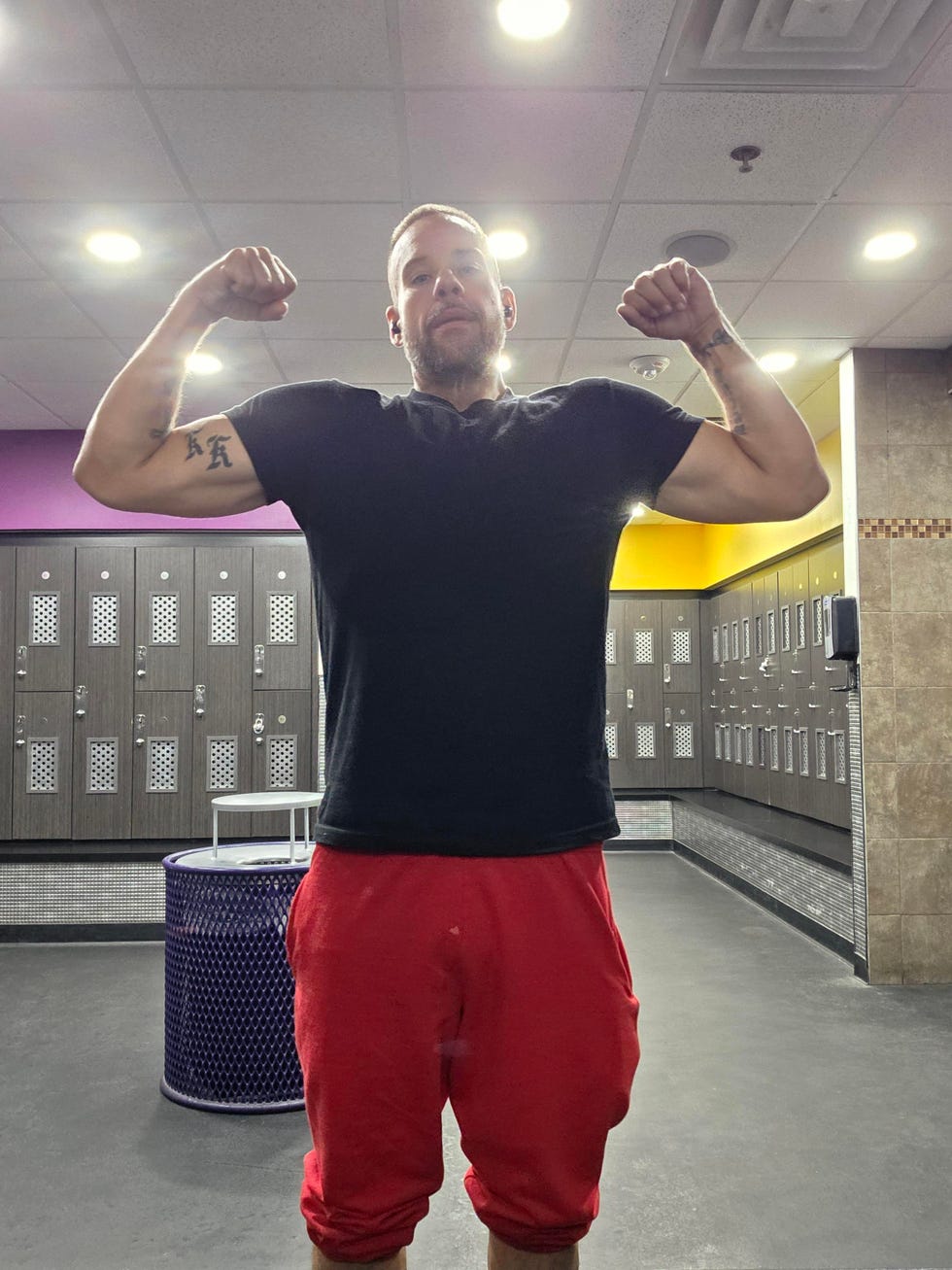 Gary Biser flexing biceps in gym locker room with bright ceiling lights.