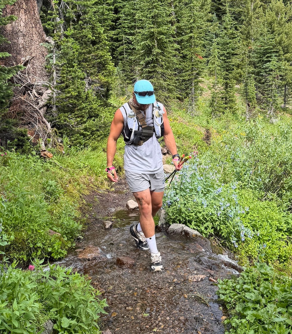 Person hiking through a forest trail with greenery and small water stream, wearing a hat and carrying trekking poles.