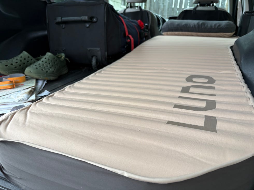 A tan camping mattress folded in the back of a vehicle, with green crocs and bags visible in the background.