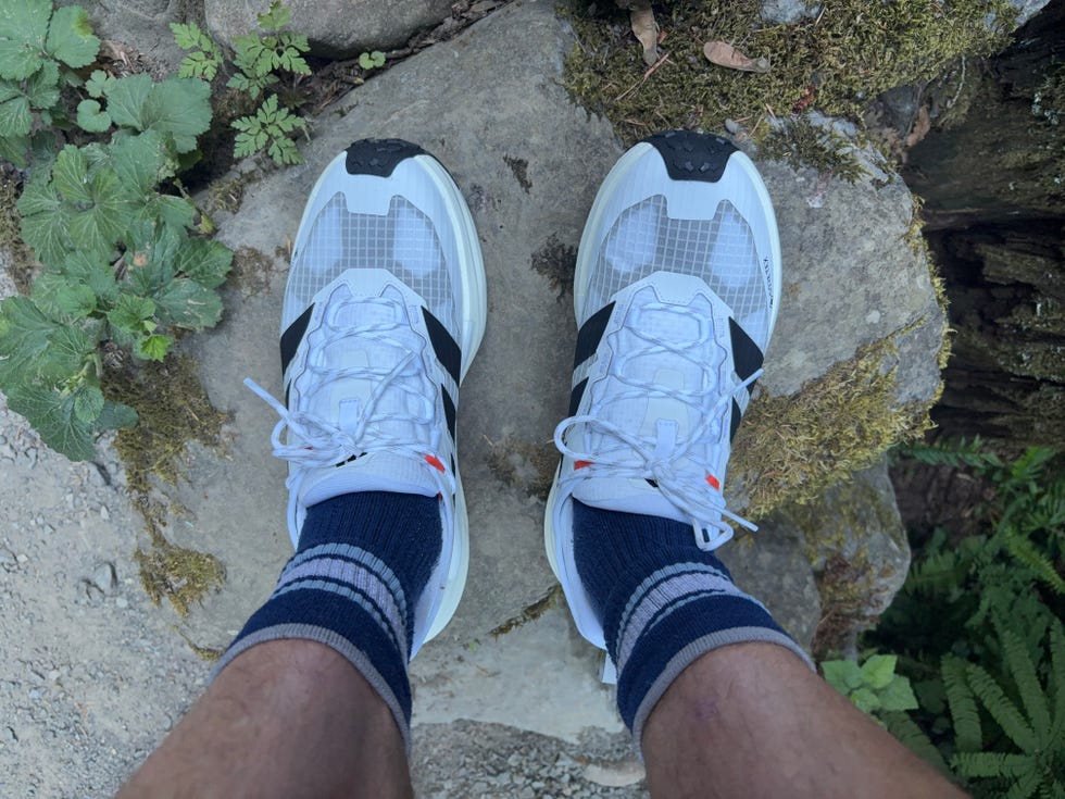 Aerial view of white athletic shoes with black stripes and blue striped socks on a rocky surface surrounded by greenery.
