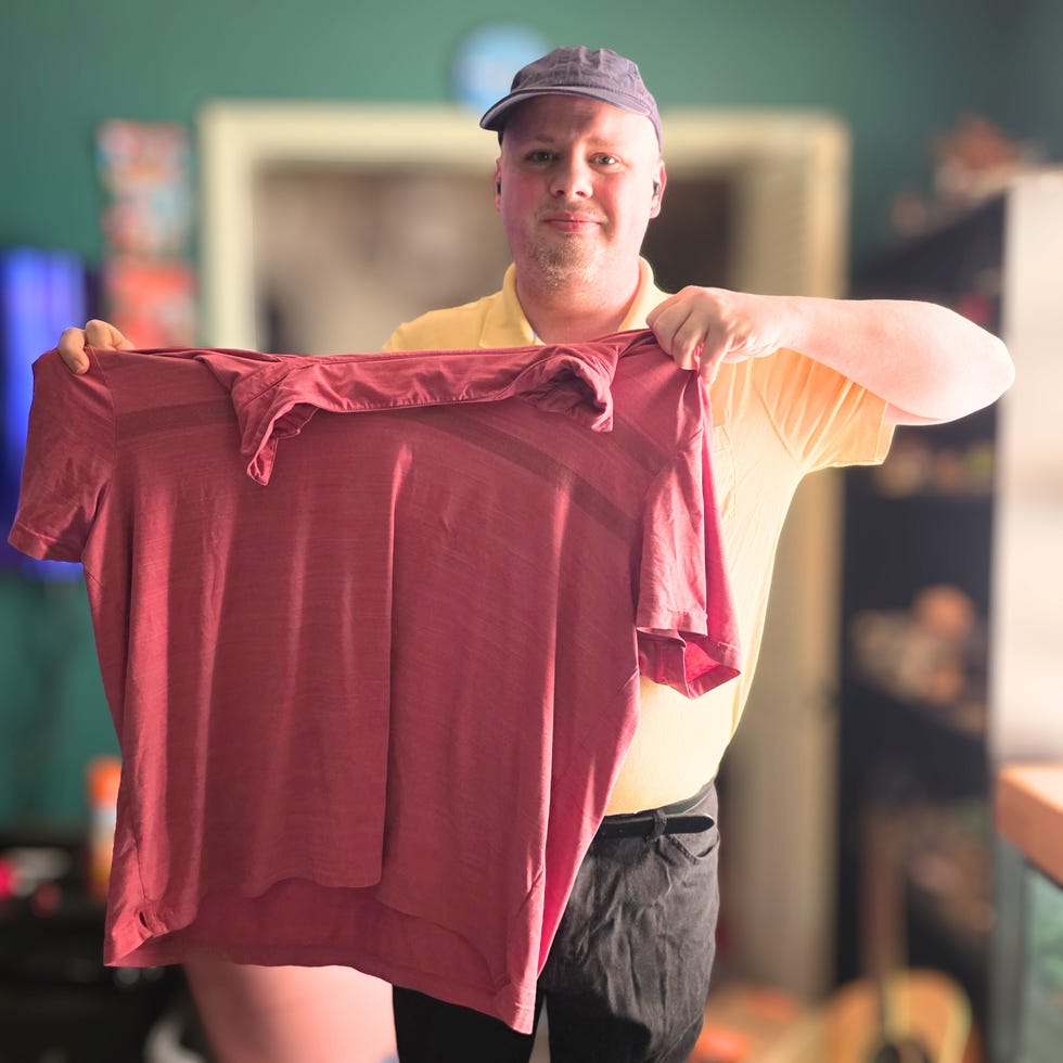 Man holding a maroon shirt indoors with a green wall and various items in the background.
