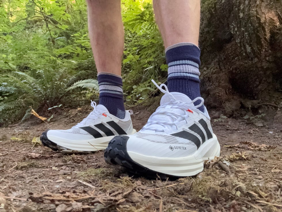 White running shoes with black stripes and orange details, worn with blue and white striped socks, in a forest setting.