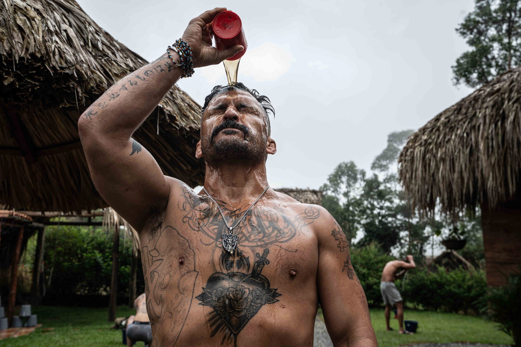 A shirtless man pours liquid over his head, showcasing numerous tattoos, in a natural outdoor setting.