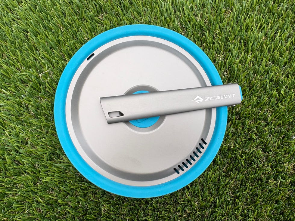 A silver camping pot lid with a blue silicone rim, placed on green grass.