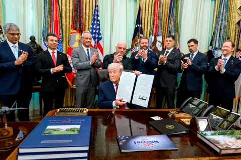 A group of officials applauds as a man holds up a signed document at a desk in an office decorated with flags.