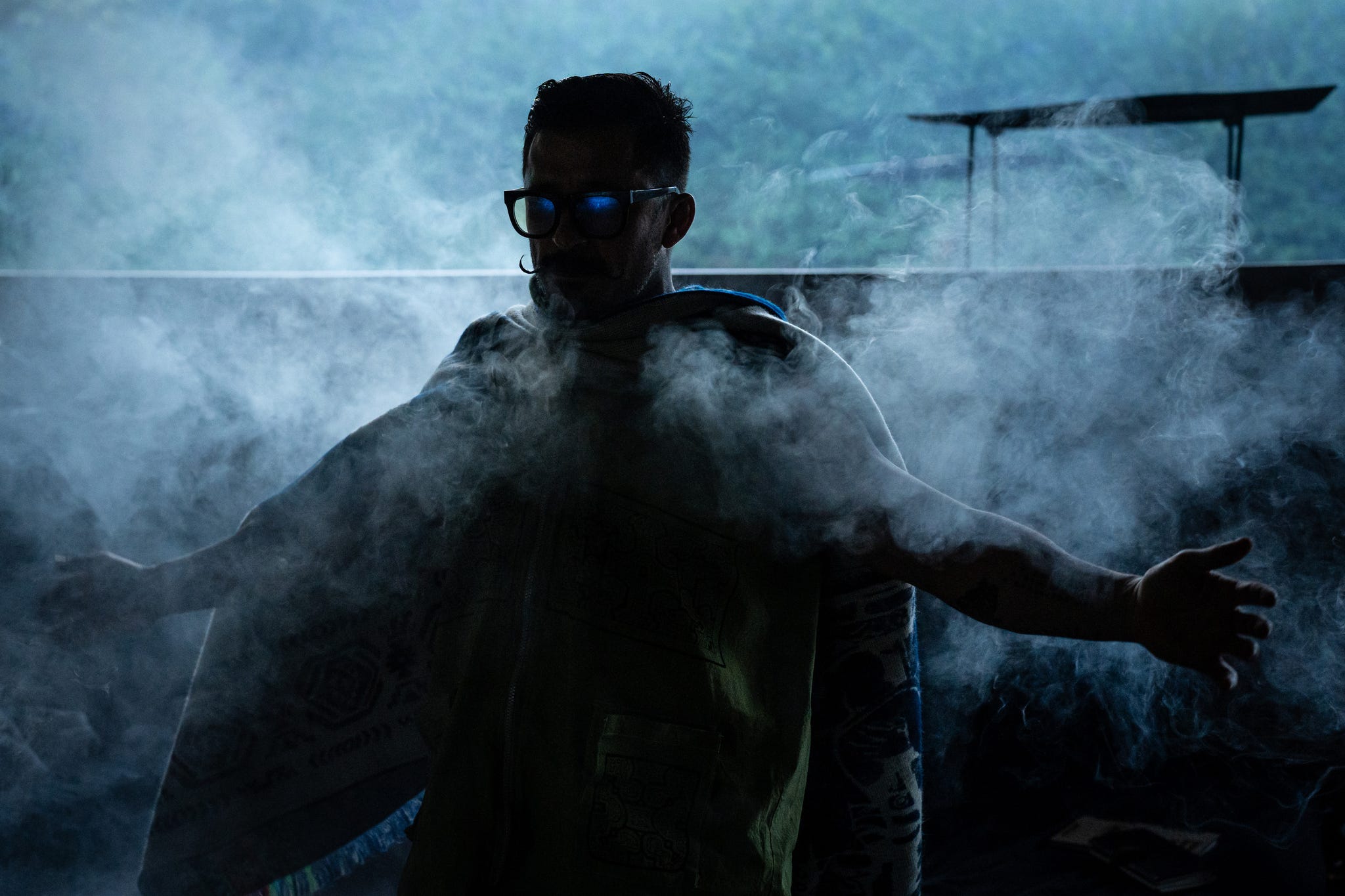 Silhouette of a figure in a poncho, wearing sunglasses, surrounded by swirling smoke in a dimly lit environment.