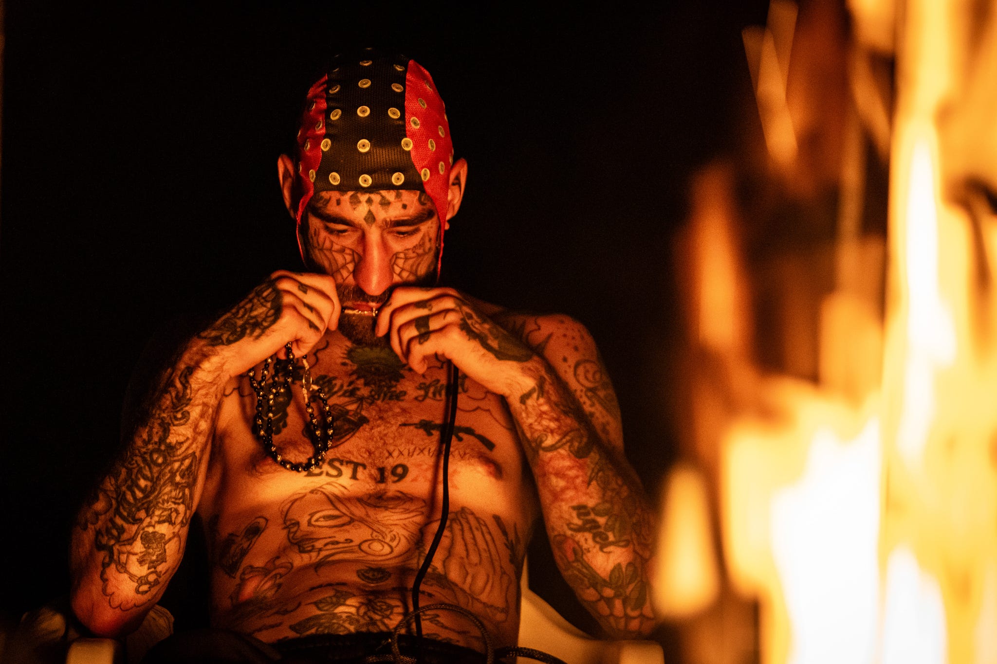 Tattooed man in a red and black bandana sits by a fire, holding beads and deep in thought.