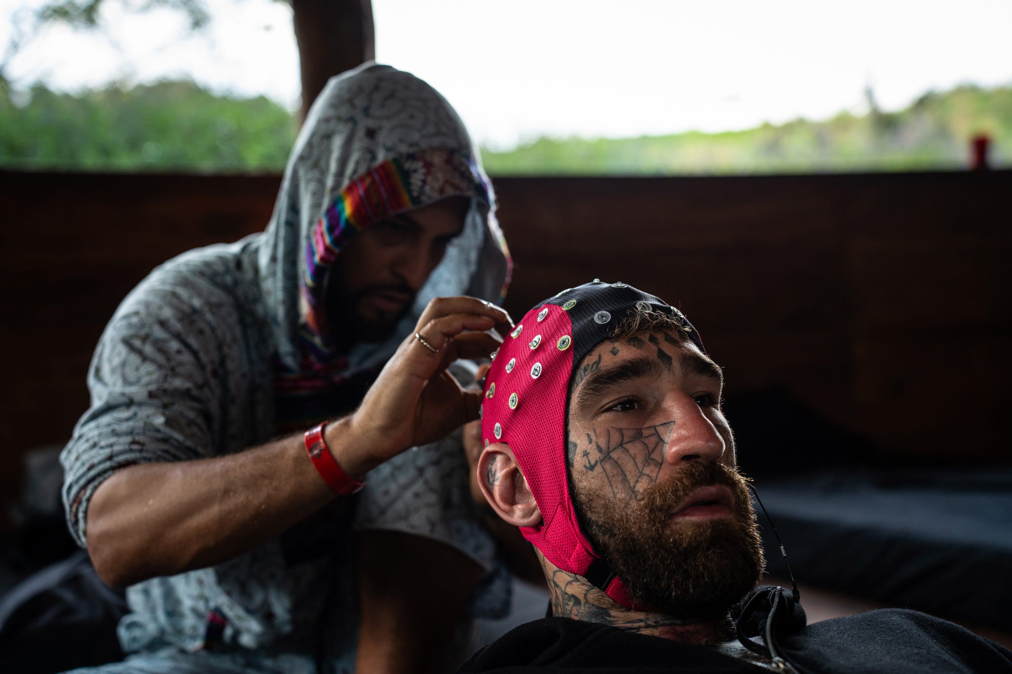A man with tattoos receives assistance in putting on an electrode cap indoors.
