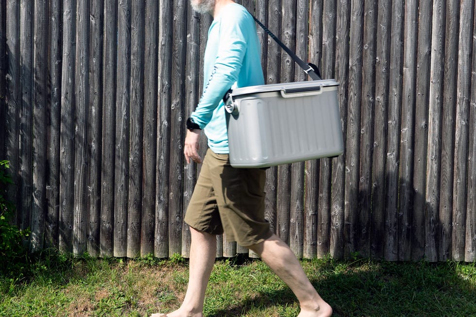 Man walking barefoot with a cooler over his shoulder beside a wooden fence on a sunny day.