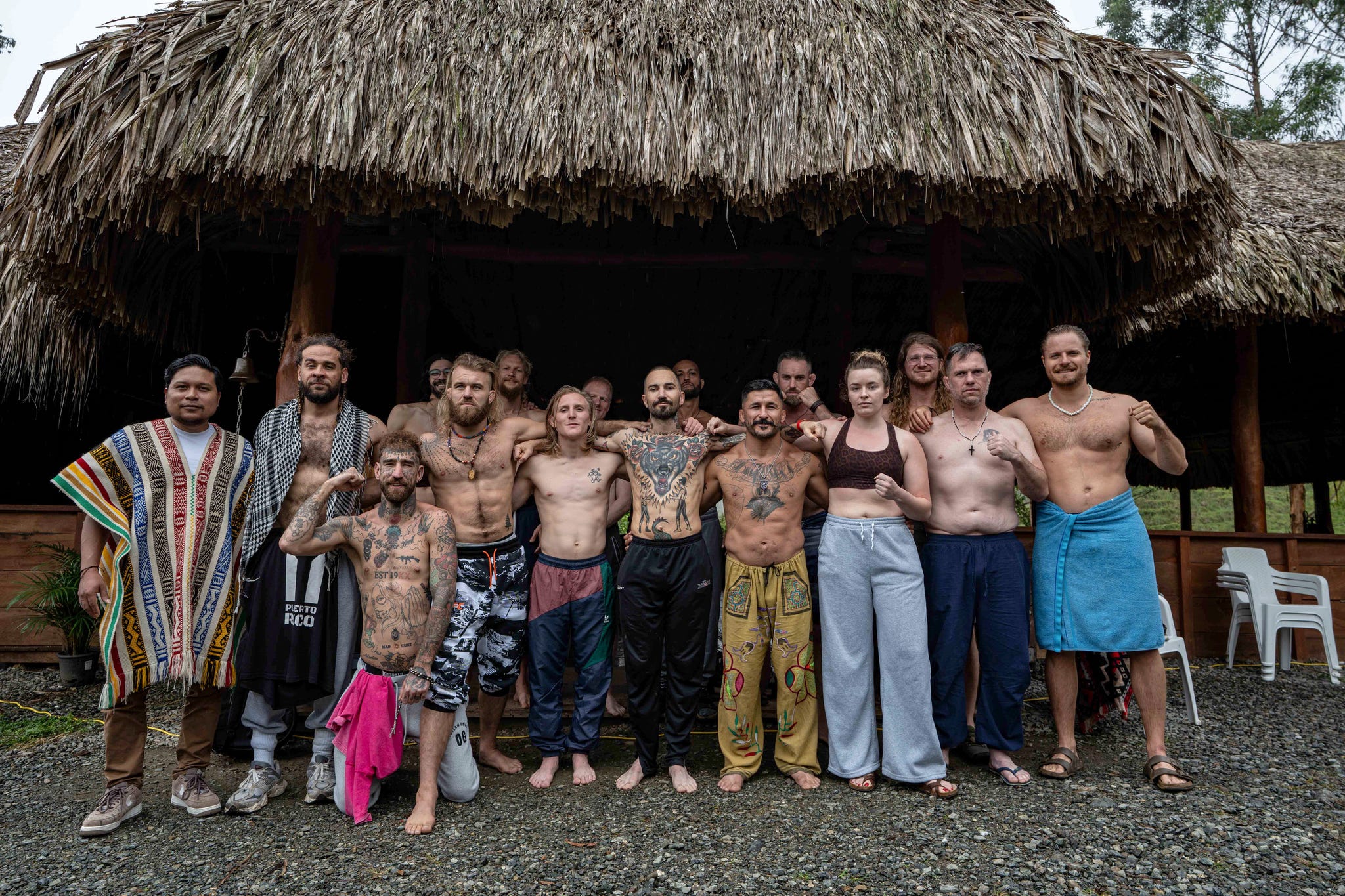 Group of people with tattoos standing together outdoors in front of a thatched-roof structure, some wearing traditional attire.