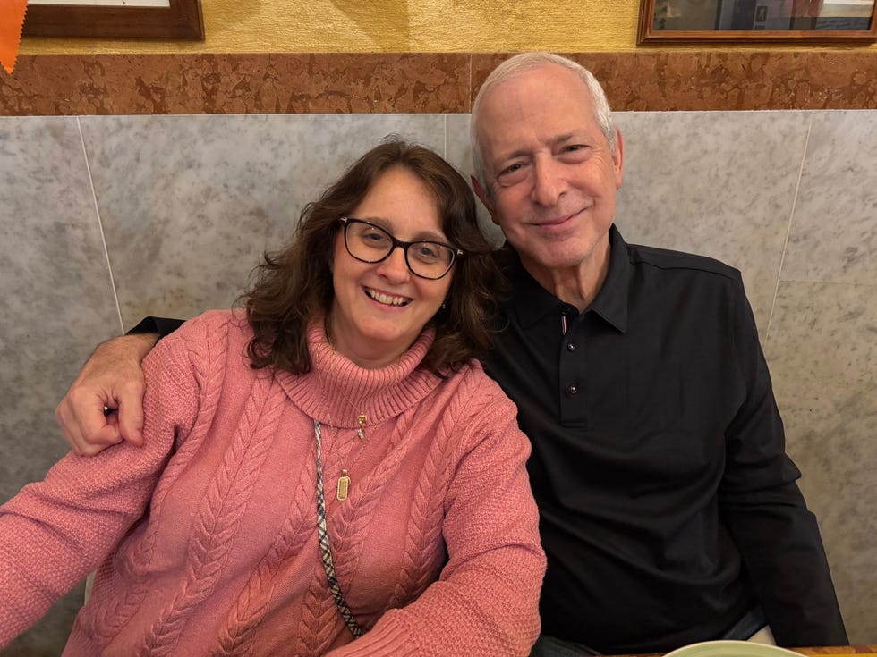 Two smiling individuals sitting closely together at a table, one wearing a pink sweater and the other in a black shirt.