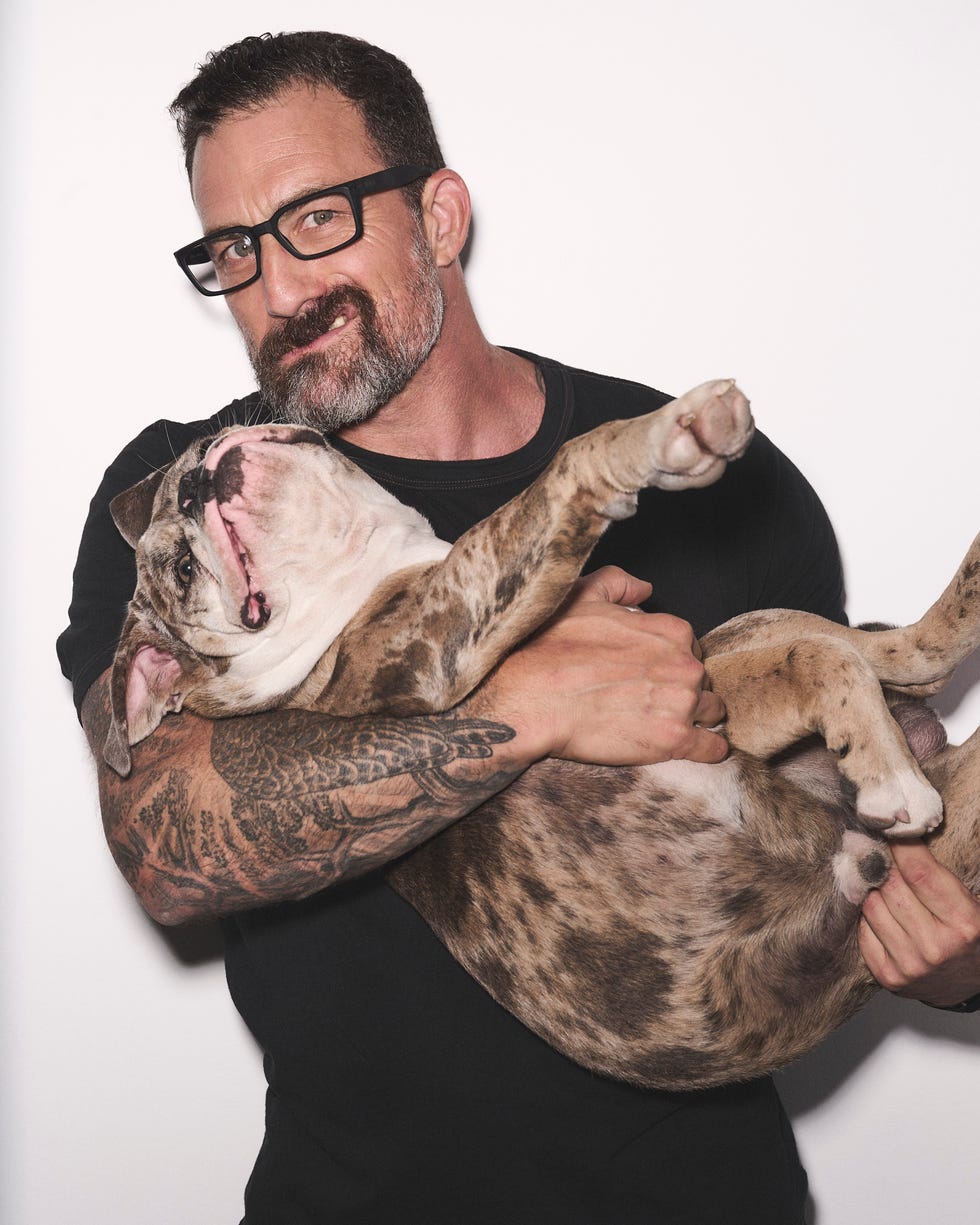 A smiling man with a beard holds a playful dog in his arms against a white background.