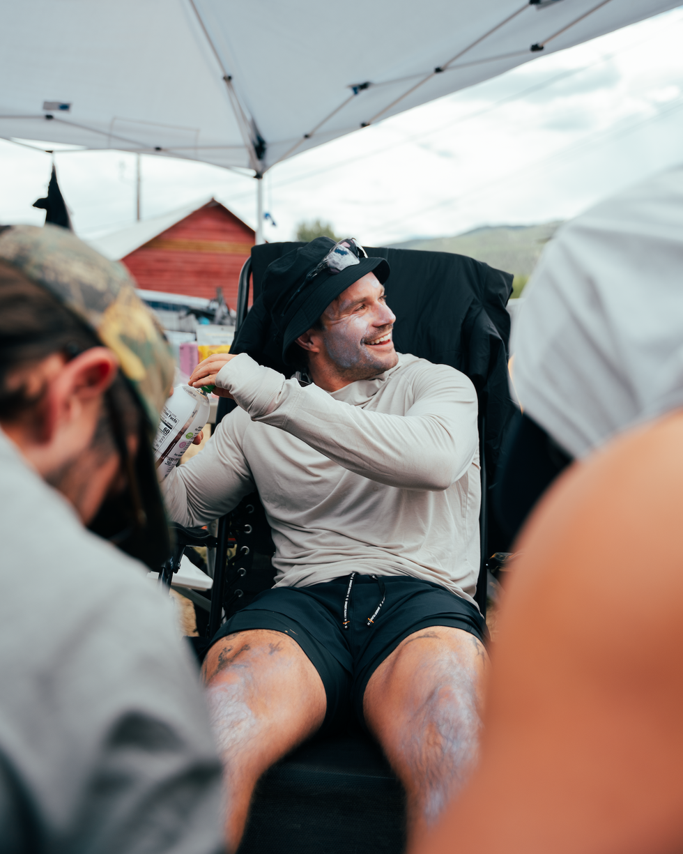 A smiling man in a hat applies sunscreen while sitting in a chair at an outdoor event, surrounded by people.