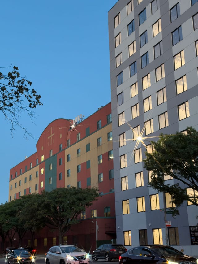 Urban building facade at dusk with colorful panels and illuminated windows, surrounded by trees and parked cars.
