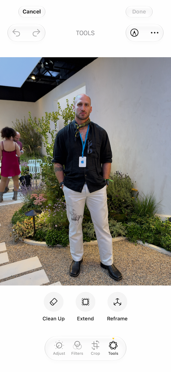 A person standing in a garden setting, wearing a black shirt and white pants with a colorful scarf and an ID badge.