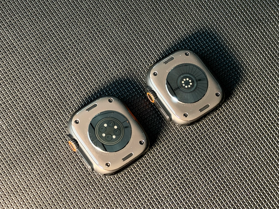 Two smartwatches viewed from below, showing sensor areas and connectors on a textured black surface.
