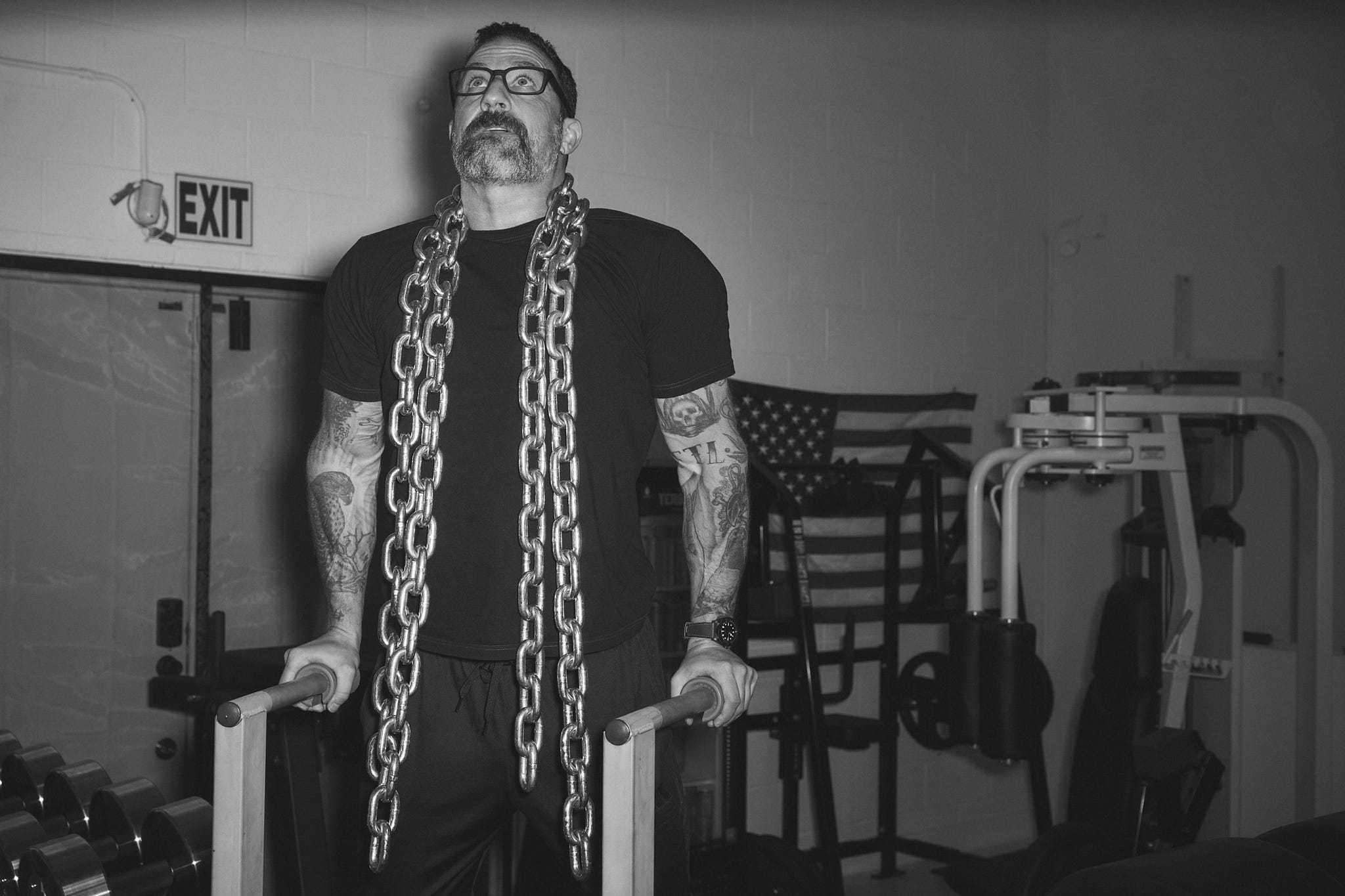 Person with tattoos wearing a chain around their neck, exercising in a gym with American flag decor.