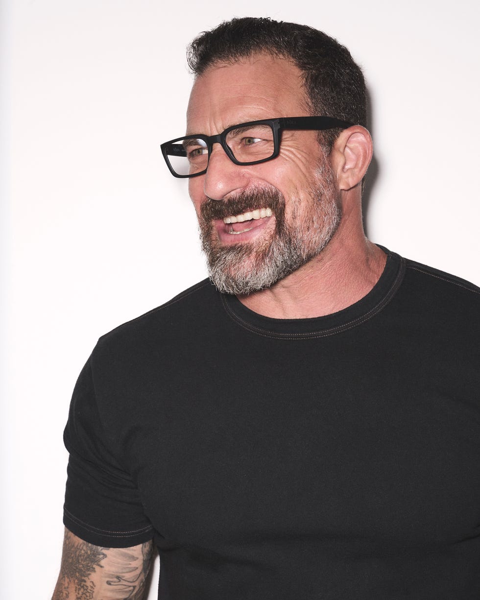 Smiling man with a beard and glasses wearing a black shirt against a light background.
