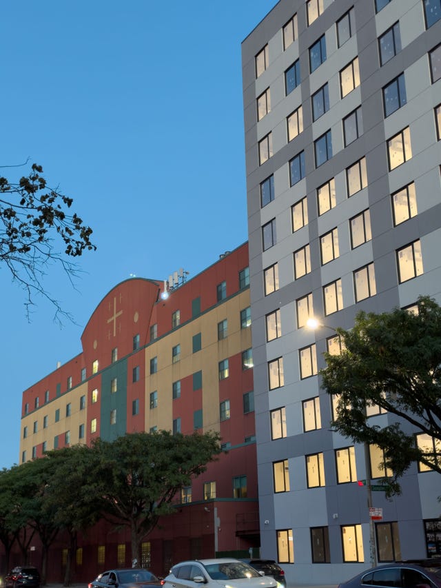 Multi-colored buildings with large windows against a clear blue evening sky, framed by trees and parked cars.
