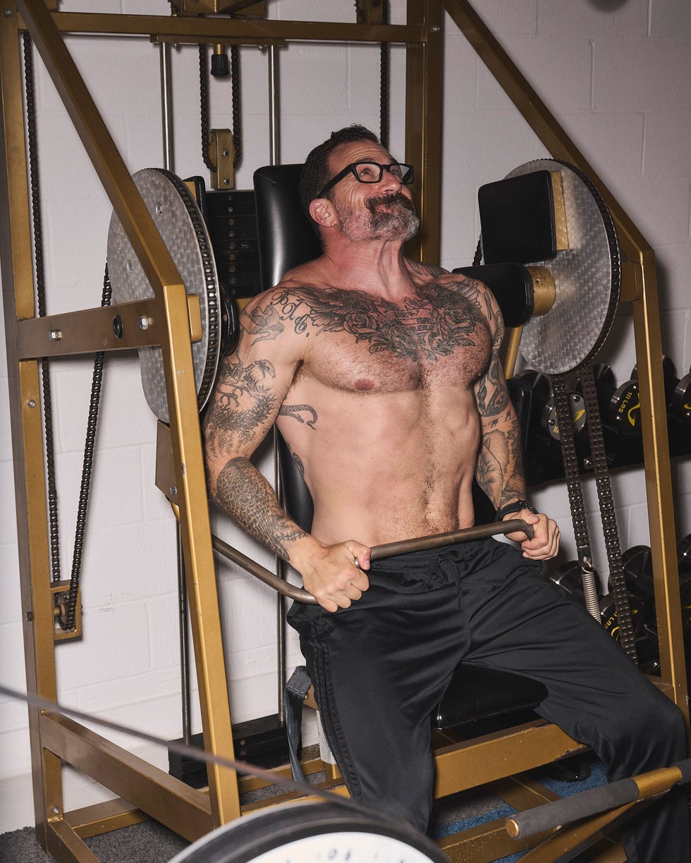 A muscular man with tattoos exercises on a weight machine in a gym setting.