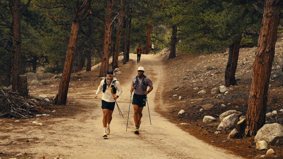 Two men running on a dirt path through a forest, with another person visible in the background.