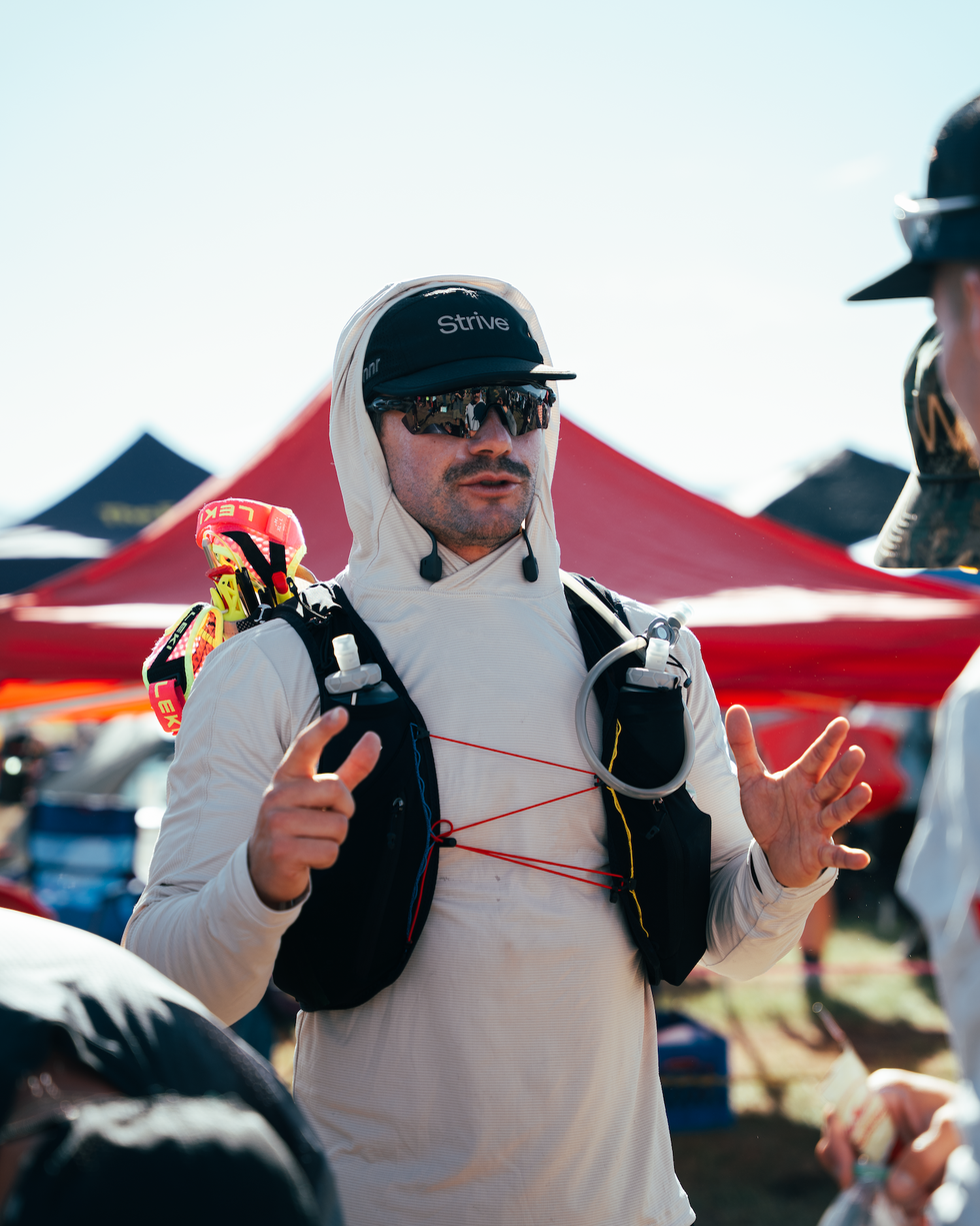 Man in athletic gear discussing at an outdoor event, wearing a cap and sunglasses with hydration pack on his chest.