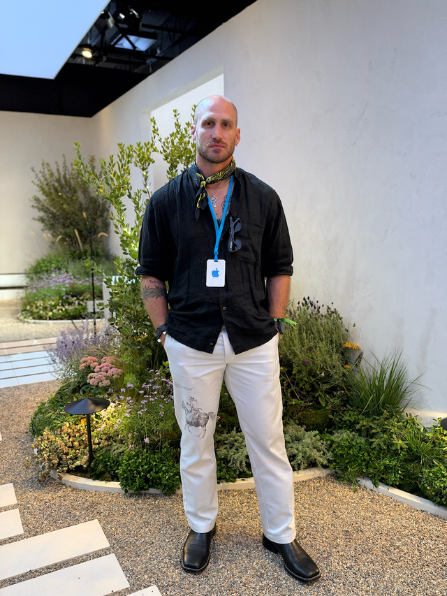 Man standing in a garden setting, wearing a black shirt, white pants with a print, and a scarf, with an event badge around his neck.