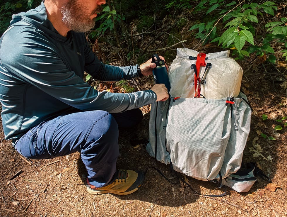 A person kneels by a backpack, preparing to secure a water bottle while surrounded by greenery on a hiking trail.