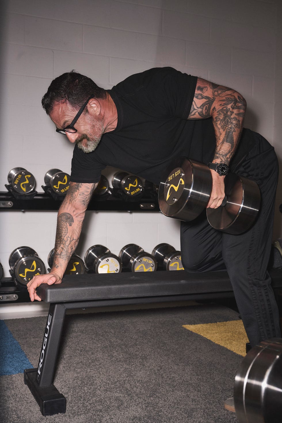 A muscular individual with tattoos lifts weights in a gym setting, focused on his exercise form.